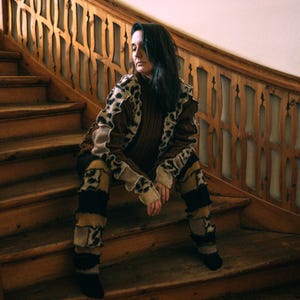 Peut inclure: Une personne porte un pull brun et un pantalon assorti avec un motif léopard. La tenue est superposée avec des accents beiges et noirs. La personne est assise sur des escaliers en bois avec une balustrade décorative.