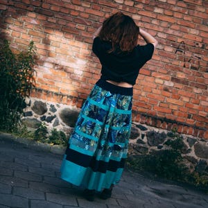 Puede incluir: Una falda larga con un estampado de hojas tropicales en tonos turquesa, azul y verde. La falda tiene rayas horizontales y se combina con una camiseta negra. La persona está de pie frente a una pared de ladrillos.