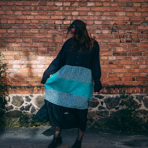 May include: A long-sleeved black dress with a color-blocked design. The dress features a teal green stripe and a floral print panel. The model is wearing black boots and standing in front of a red brick wall.