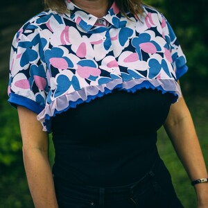 May include: A cropped button-up shirt with a floral pattern in blue and pink on a white background. The shirt has a blue collar and cuffs, and a ruffled trim. The shirt is worn over a black top.