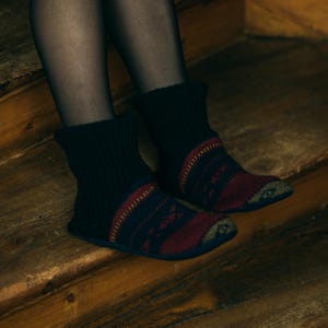 May include: Black knitted ankle boots with a red and navy blue pattern. The boots have a flat sole and are worn on a wooden surface. The boots have a warm, cosy appearance, perfect for indoor wear.