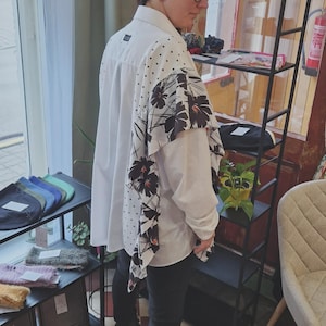 Può includere: Camicia bianca con un design floreale e a pois unico sul retro e sulle maniche. Il motivo floreale presenta fiori neri con centri marroni. La camicia è abbinata a pantaloni neri, creando un contrasto elegante.