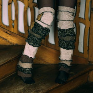 May include: A pair of leg warmers in shades of beige, brown, and olive green. The leg warmers have a layered design with ruffled edges and are worn over black tights. The image is taken indoors, with a wooden background.