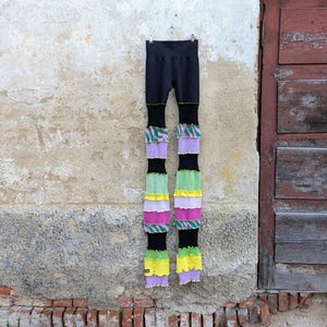 Op de afbeelding: Zwarte legging met een hoge taille en lange, veelkleurige, geribbelde beenverlengingen. De verlengingen bevatten horizontale banden in groen, geel, paars, roze en een groen-roze zebra-achtig patroon met golvende randen.