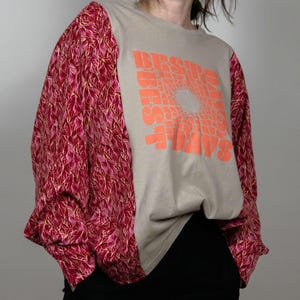 Könnte beinhalten: Ein beiges Langarmshirt mit einem grafischen Druck der Worte "BE SURE TO" in einem kreisförmigen Muster. Die Ärmel sind aus einem roten und rosafarbenen Blumenstoff.