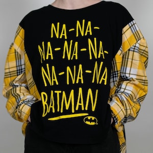 May include: Black t-shirt with yellow text that reads "NA-NA-NA-NA-NA BATMAN" and a yellow Batman logo. The shirt has yellow and black plaid sleeves.