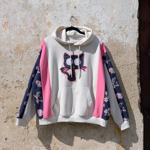 Könnte beinhalten: Ein cremefarbener Kapuzenpullover mit einer zentralen schwarzen Katzengrafik mit großen weißen Augen und rosa Akzenten. Das Kleidungsstück verfügt über eine Kängurutasche, leuchtend rosa Seitenteile und lange Ärmel mit dunkelblauen Blumenmustern.