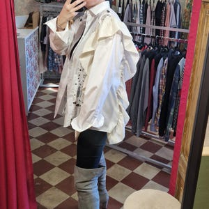 Puede incluir: Camisa blanca abotonada con volantes y diseños abstractos negros. La camisa se combina con leggings negros y botas altas grises. La imagen está tomada en una tienda de ropa, con percheros de ropa visibles en el fondo.