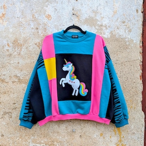May include: A colour-blocked sweatshirt featuring a central black panel with a white unicorn graphic, accented by bright pink, teal, and yellow sections. The sleeves have black and teal zebra-like stripes, and the interior label reads RE.MELIN.