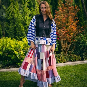Op de afbeelding: Een vrouw draagt een patchwork rok en top. De top heeft blauw-wit gestreepte mouwen, een zwart lijfje en bloemen- en kanten details. De rok is een mix van stoffen met patronen in rood, roze, paars en wit. De achtergrond is een groene tuin.