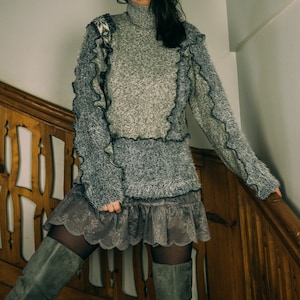 Op de afbeelding: Een grijze trui met een hoge hals en ruches. De trui heeft een textuur en wordt gecombineerd met een grijze kanten rok. De outfit wordt gecompleteerd met grijze suède laarzen, waardoor een gelaagde, getextureerde look ontstaat.