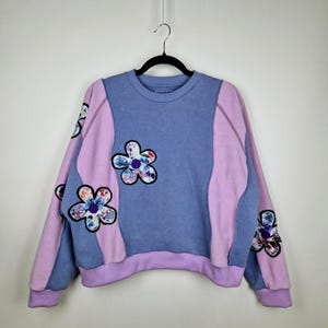 Può includere: Una felpa girocollo nei toni del blu e lavanda, ornata da applicazioni floreali. La felpa presenta un design a blocchi di colore con maniche e pannelli laterali lavanda e toppe floreali.