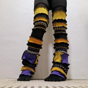 Puede incluir: Calentadores de piernas multicolores en negro, amarillo y morado con volantes decorativos y secciones estampadas. Los calentadores se usan sobre leggings negros y presentan una mezcla de texturas y colores.