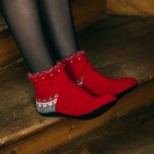 Puede incluir: Un par de calcetines tobilleros rojos de punto con un diseño decorativo en blanco y gris alrededor del puño y el talón. Los calcetines tienen suelas negras y se llevan sobre medias negras. Los calcetines están sobre una superficie de madera.