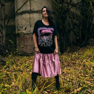 Può includere: Una maglietta nera con scollo a V con la scritta "INTENSE & SMOOTH" e una grafica di labbra rosa. La modella indossa una gonna midi in raso rosa e collant neri. Lo sfondo comprende alberi e una struttura in cemento.