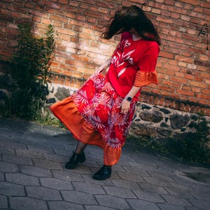 Peut inclure: Une robe rouge avec un motif floral blanc et un haut rouge assorti avec des accents floraux blancs. La robe a une jupe superposée avec une sous-couche orange et un motif de feuilles de palmier. La tenue est associée à des bottes noires.