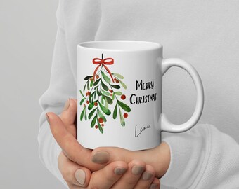 May include: White ceramic mug featuring a watercolor design of mistletoe with green leaves, red berries, and a red bow. The mug has the text "Merry Christmas" and the name "Luna".