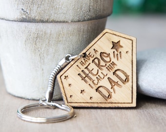 May include: Wooden keychain with a superhero design and the text "I have a hero I call him Dad".