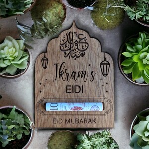 Op de afbeelding: Een houten Eid Mubarak geldhouder met een gleuf voor contant geld. De houder is versierd met Arabische kalligrafie, sterren en een lantaarnontwerp. Er staat "Ikram's Eidi" en "Eid Mubarak" op.