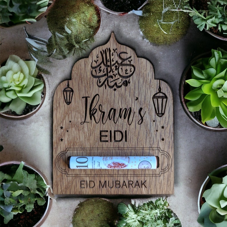 Op de afbeelding: Een houten Eid Mubarak geldhouder met een gleuf voor contant geld. De houder is versierd met Arabische kalligrafie, sterren en een lantaarnontwerp. Er staat "Ikram's Eidi" en "Eid Mubarak" op.