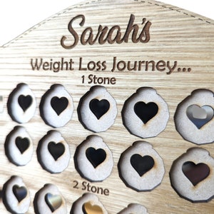 May include: Wooden weight loss tracker with the name "Sarah's Weight Loss Journey..." and the text "1 Stone" and "2 Stone". The tracker has a series of heart-shaped cutouts, some of which are filled with gold hearts.