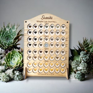 May include: A wooden weight loss tracker with the name "Sarah's Weight Loss Journey..." engraved on it. The tracker has 56 heart-shaped slots, with the first 7 slots labelled "1 Stone", the next 14 slots labelled "2 Stone", the next 14 slots labelled "3 Stone", and the final 21 slots labelled "4 Stone". The tracker also has a label that reads "one pound at a time".