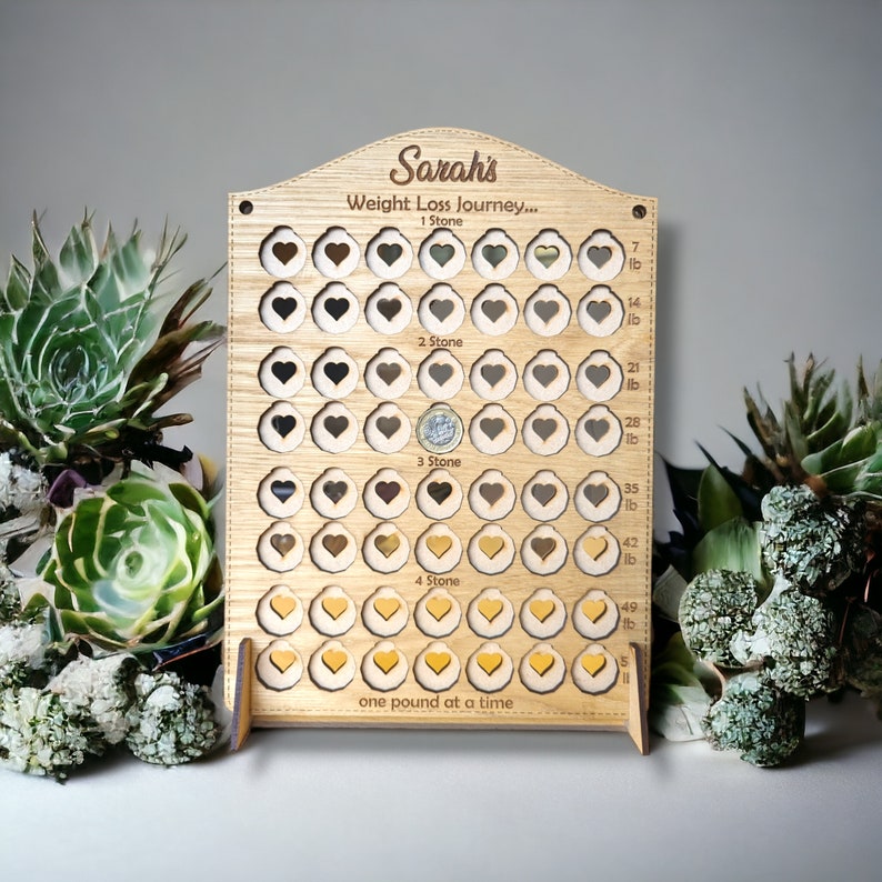 May include: A wooden weight loss tracker with the name "Sarah's Weight Loss Journey..." engraved on it. The tracker has 56 heart-shaped slots, with the first 7 slots labelled "1 Stone", the next 14 slots labelled "2 Stone", the next 14 slots labelled "3 Stone", and the final 21 slots labelled "4 Stone". The tracker also has a label that reads "one pound at a time".