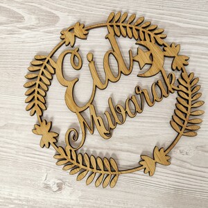 May include: Wooden cutout of a wreath with the words "Eid Mubarak" in a decorative font. The wreath is made of leaves and flowers.