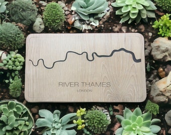 May include: A wooden chopping board with a black outline of the River Thames and the text "River Thames, London". The board is surrounded by green succulents.