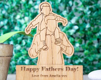May include: A wooden Father's Day decoration featuring a father carrying a child on his shoulders. The base reads "Happy Fathers Day! Love from Amelia xxx". The wooden figure is light brown and stands on a small wooden base. The background is a blurred green.