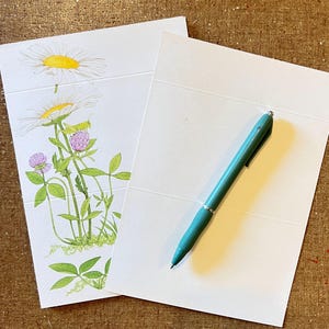 Puede incluir: Dos tarjetas de felicitación blancas con líneas en relieve, una con una ilustración en acuarela de margaritas, tréboles y un saltamontes. Un bolígrafo turquesa descansa sobre la otra tarjeta. El fondo es una superficie marrón texturizada.