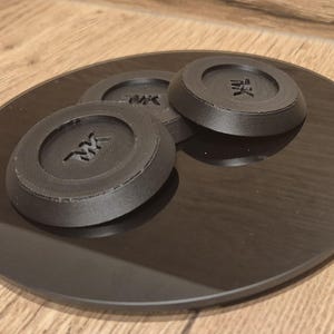 Könnte beinhalten: Drei schwarze, runde, scheibenförmige Objekte mit dem Wort "MK" auf jedem. Die Objekte befinden sich auf einer schwarzen Oberfläche.