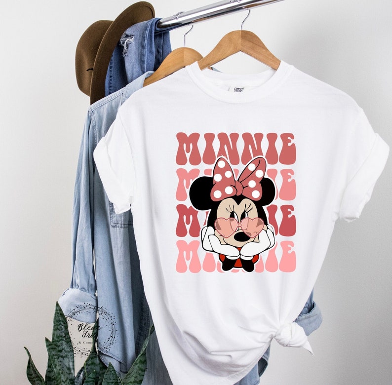 K&ouml;nnte beinhalten: Wei&szlig;es T-Shirt mit einem pinkfarbenen und roten Aufdruck von Minnie Maus mit Sonnenbrille und roter Schleife. Der Text "MINNIE" wird in einer Retro-Schrift wiederholt.