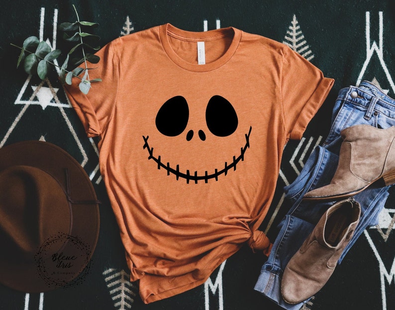 Puede incluir: Una camiseta naranja quemada con un gr&aacute;fico de cara de calabaza negra inspirado en Jack Skellington. La camiseta est&aacute; colocada sobre una superficie con estampado blanco y negro.