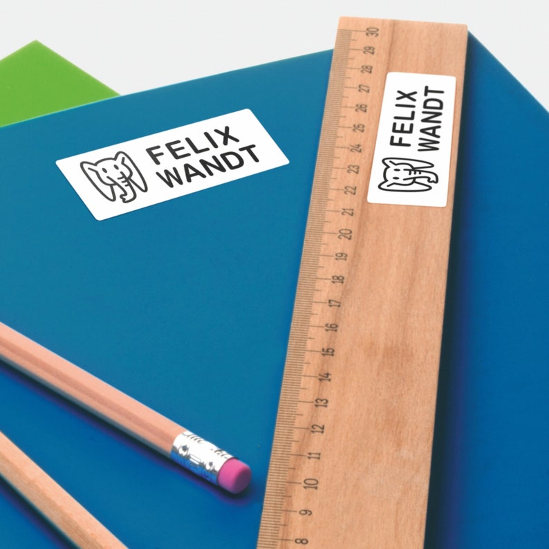 May include: Two white rectangular labels with black text and an elephant illustration. The labels read "Felix Wandt". The labels are on a blue notebook and a wooden ruler.