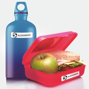 May include: A red lunchbox with a green apple and a sandwich inside. The lunchbox has a sticker with a soccer ball and the name "ALEXANDER" on it. A blue water bottle with a sticker with a soccer ball and the name "ALEXANDER" on it is next to the lunchbox.