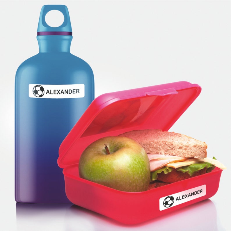 May include: A red lunchbox with a green apple and a sandwich inside. The lunchbox has a sticker with a soccer ball and the name "ALEXANDER" on it. A blue water bottle with a sticker with a soccer ball and the name "ALEXANDER" on it is next to the lunchbox.