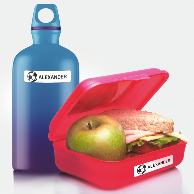 May include: A red lunchbox with a green apple and a sandwich inside. The lunchbox has a sticker with a soccer ball and the name "ALEXANDER" on it. A blue water bottle with a sticker with a soccer ball and the name "ALEXANDER" on it is next to the lunchbox.