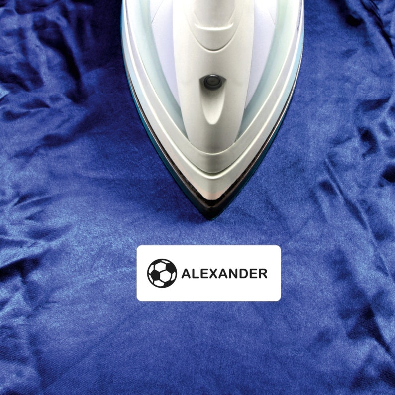 May include: A white iron with a black cord is pressing down on a blue fabric with a white iron-on label that says "ALEXANDER" with a black soccer ball icon.