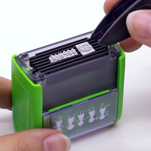 May include: A green and gray self-inking stamp with a clear plastic top. The stamp has a cat design and the text "BOGHIE" on the top. The stamp is being held by a hand with tweezers.
