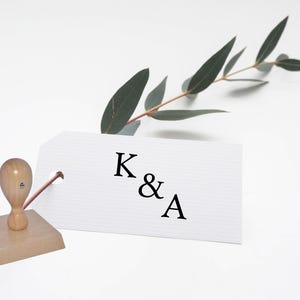 May include: A wooden stamp with a handle and a square base. The stamp is used to create an imprint of the letters "K & A" on a white card with a sprig of greenery.