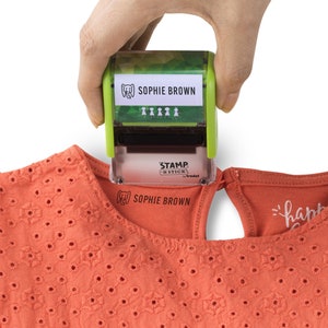 May include: A green and white self-inking stamp with the text "SOPHIE BROWN" and a small elephant icon. The stamp is being used to stamp the text "SOPHIE BROWN" onto a peach colored fabric.