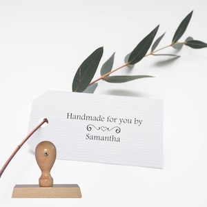 May include: A wooden stamp with a light brown handle and base, beside a white tag with the text "Handmade for you by Samantha." A sprig of green leaves is behind the tag. The stamp is likely used for crafting or personalising items.
