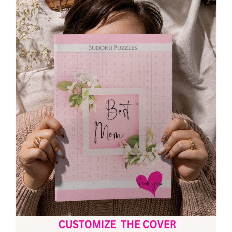 May include: A pink and white book with a floral design and the text "Best Mom" in a white frame. The book has a Sudoku puzzle grid on the cover and the text "Love you" in a pink heart.