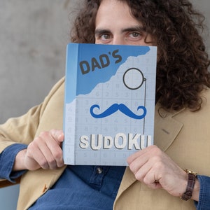 Pu&ograve; includere: Un libro blu con il testo "DAD'S SUDOKU" sulla copertina. Il libro ha un sudoku in bianco e nero sulla copertina con un'immagine grafica di baffi neri e monoccolo.