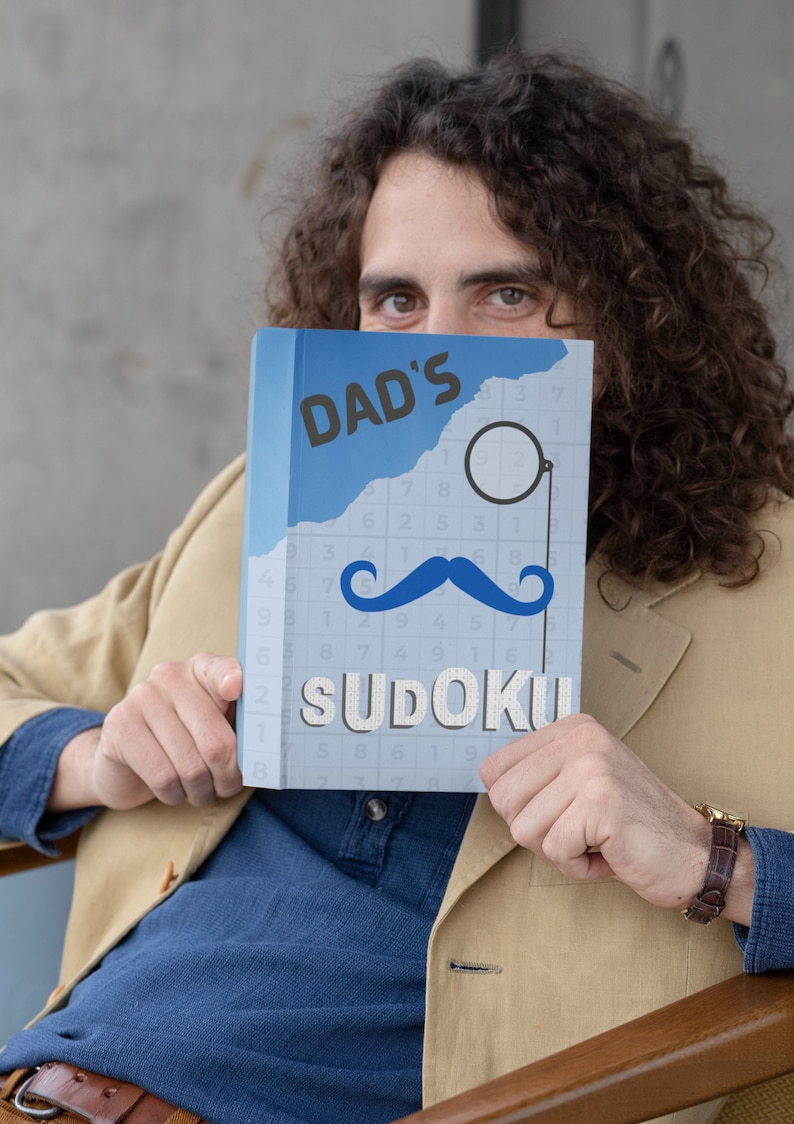 Pu&ograve; includere: Un libro blu con il testo "DAD'S SUDOKU" sulla copertina. Il libro ha un sudoku in bianco e nero sulla copertina con un'immagine grafica di baffi neri e monoccolo.