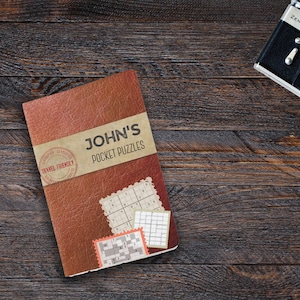Może przedstawiać: Brązowa książka w sk&oacute;rzanej oprawie z tytułem "JOHN'S POCKET PUZZLES" i stemplem "TRAVEL FRIENDLY". Książka leży na drewnianej powierzchni, obok niej znajdują się aparat fotograficzny i okulary.