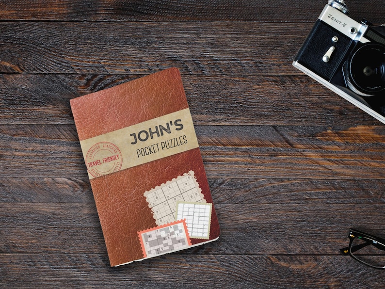 Może przedstawiać: Brązowa książka w sk&oacute;rzanej oprawie z tytułem "JOHN'S POCKET PUZZLES" i stemplem "TRAVEL FRIENDLY". Książka leży na drewnianej powierzchni, obok niej znajdują się aparat fotograficzny i okulary.