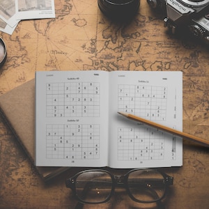 Może przedstawiać: Otwarta książka z dwoma łamigł&oacute;wkami Sudoku, jedna oznaczona jako "Sudoku 49", a druga jako "Sudoku 50". Oł&oacute;wek leży na książce, a na stole leży lupa, aparat fotograficzny i okulary.