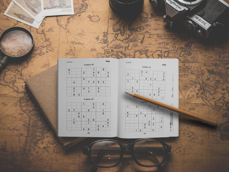 Może przedstawiać: Otwarta książka z dwoma łamigł&oacute;wkami Sudoku, jedna oznaczona jako "Sudoku 49", a druga jako "Sudoku 50". Oł&oacute;wek leży na książce, a na stole leży lupa, aparat fotograficzny i okulary.